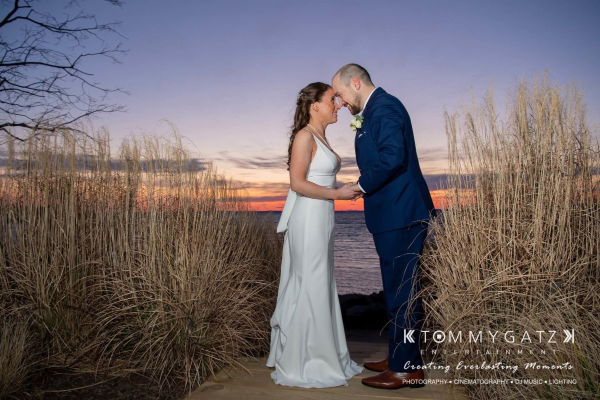 A couple in wedding attire stands close together, holding hands, with a sunset over a body of water in the background.