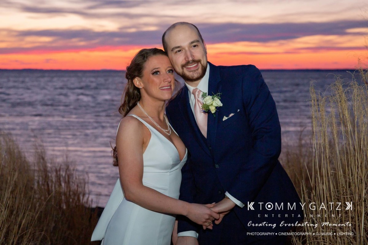 A couple in formal attire poses by a lake during sunset, surrounded by tall grass.
