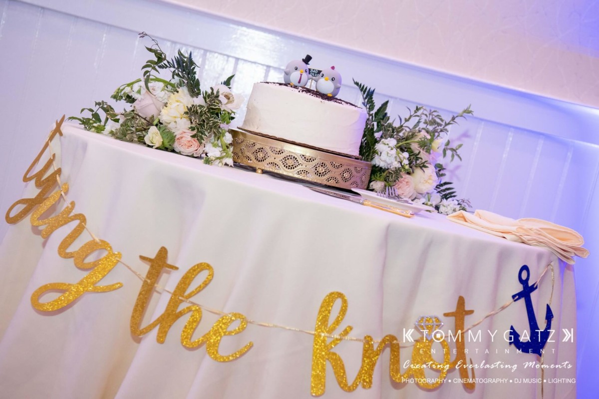 Wedding cake on a decorated table with floral arrangements and a "tying the knot" sign.