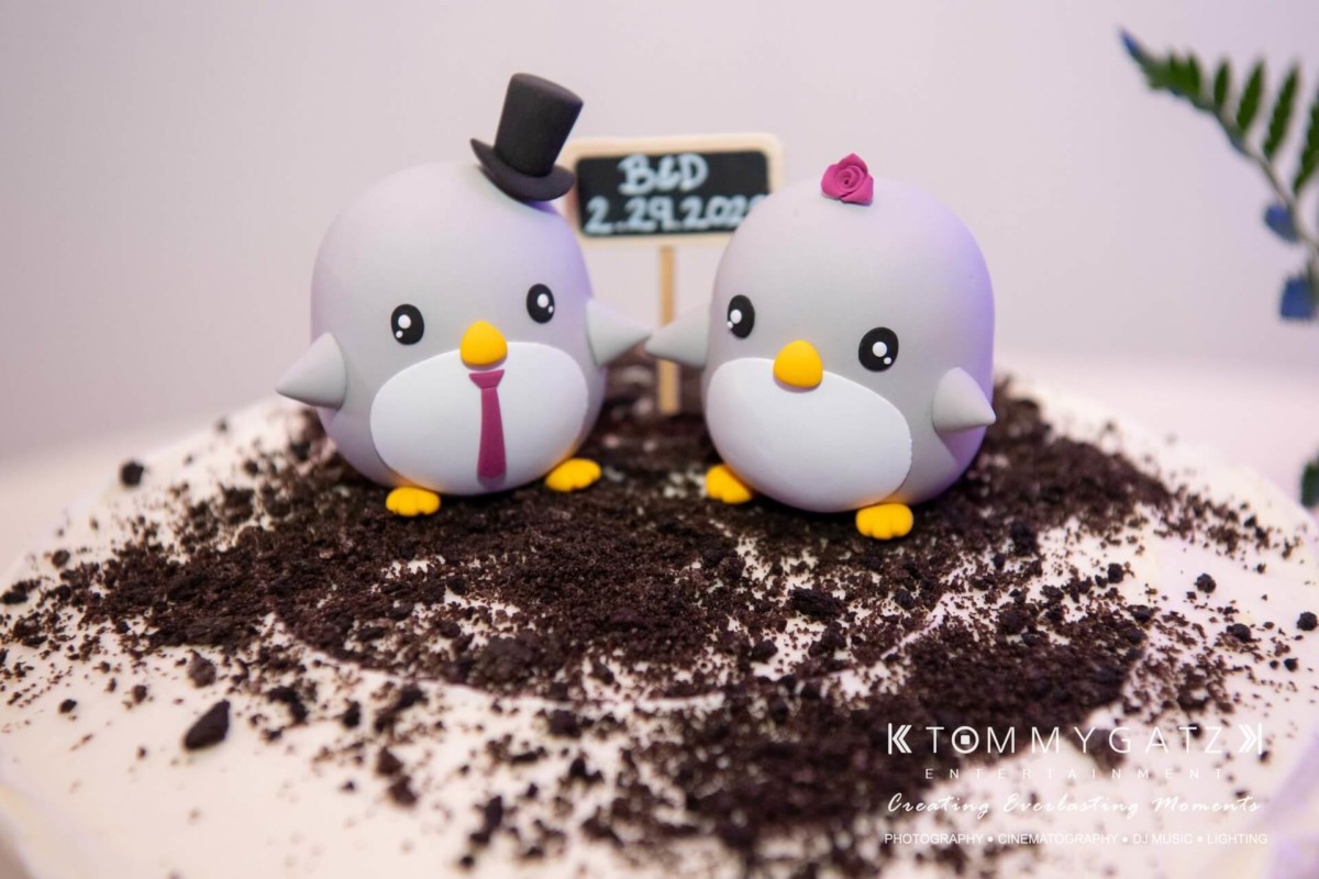 Two penguin cake toppers on a dirt-like surface; one wears a top hat, the other has a pink flower. A small sign reads "BLD 2.29.2020.