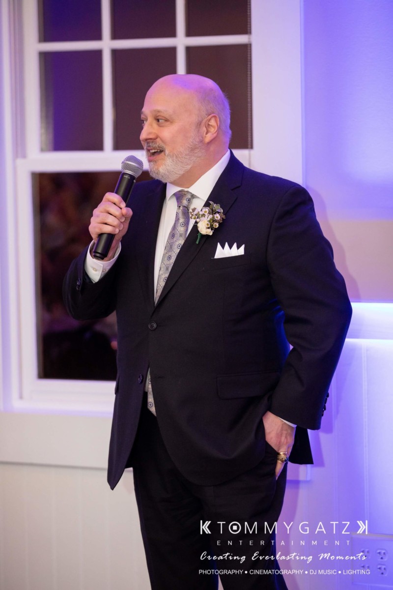 A man in a suit speaks into a microphone at an indoor event.