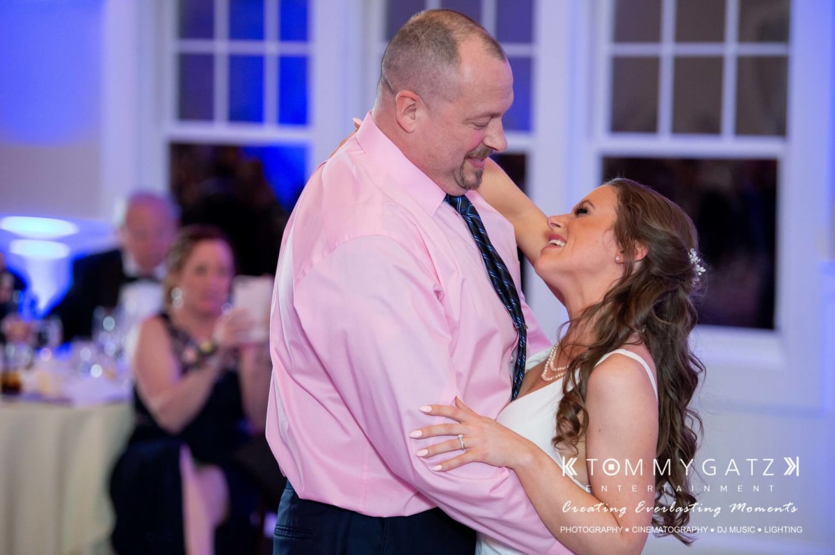 A couple dances and smiles at an indoor event, with guests seated in the background.