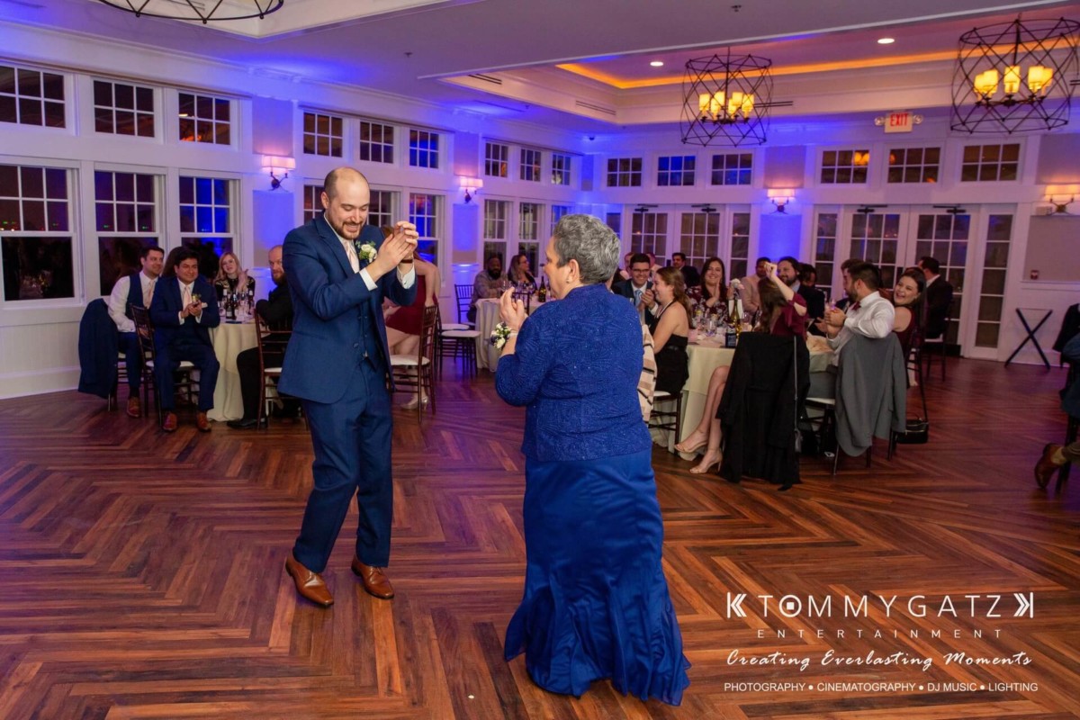 A man and woman in formal attire are dancing joyfully on a wooden floor in a banquet hall, with guests seated at tables in the background.