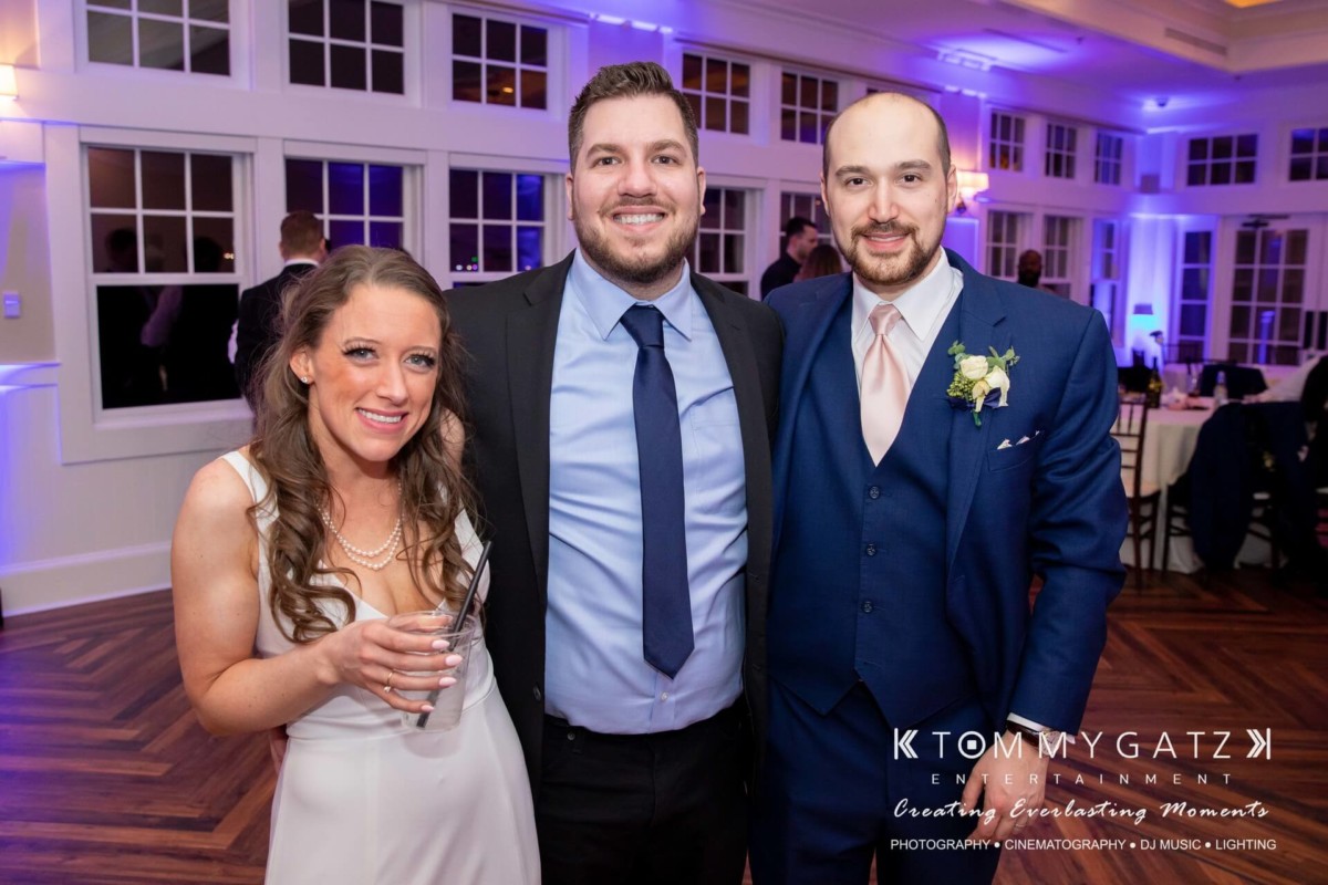 Three people pose together at an indoor event. The woman on the left holds a drink, while both men on the right wear suits.