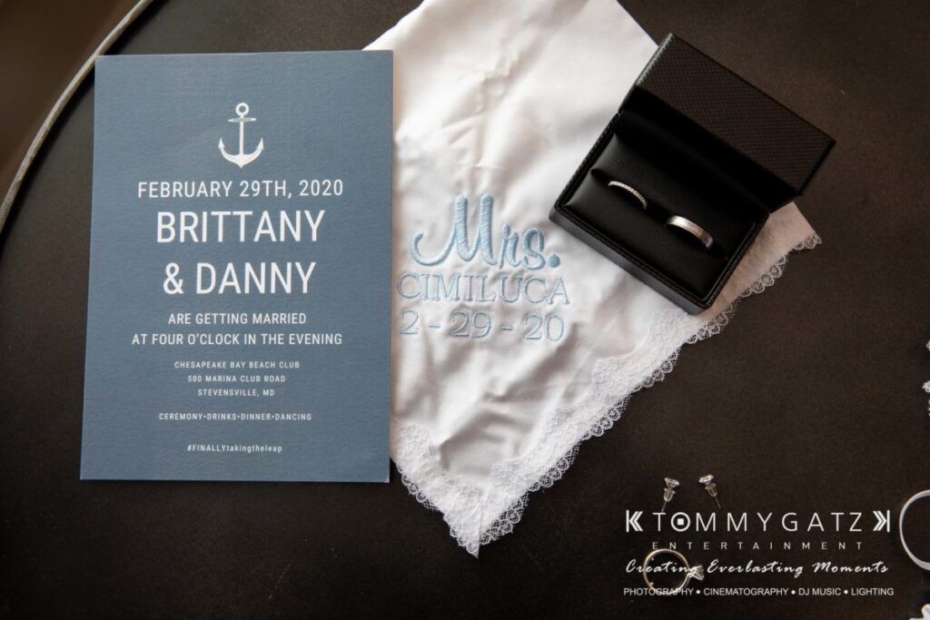 Wedding invitation and accessories including a handkerchief with "Mrs. Cimiluca 2-29-20" embroidered, two rings in a box, and promotional materials for Tommy Gatz Entertainment.