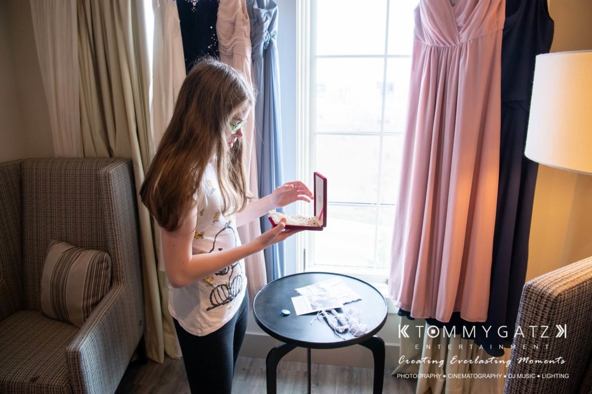 A young woman stands by a window, holding an open jewelry box. Dresses hang nearby. The scene appears to be part of a preparation or dressing area.