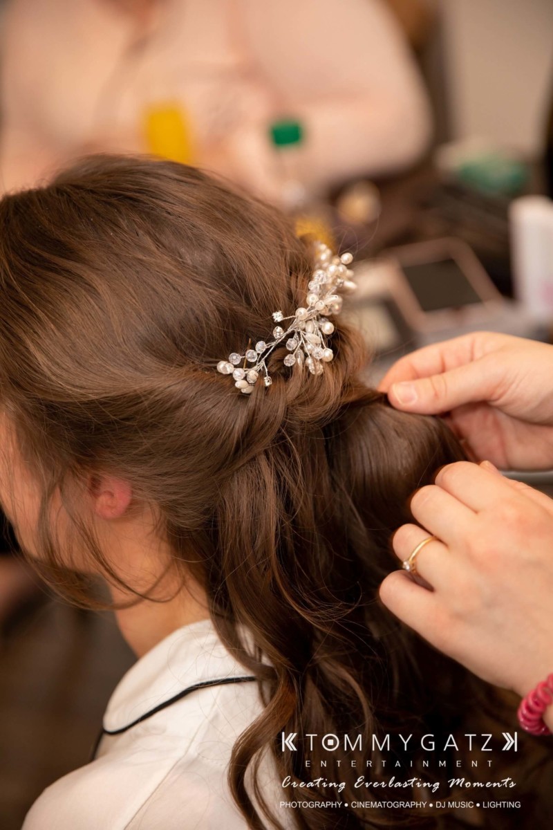 Person styling a woman's hair, adorned with a decorative hairpiece, during a formal preparation.