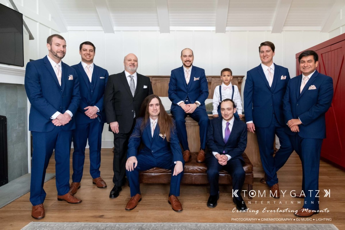 A group of nine men in formal attire pose indoors, some seated and others standing. The setting includes a fireplace and red barn-style doors.