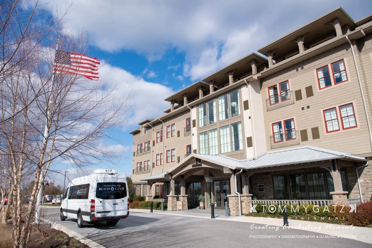 A hotel with a large entrance and an American flag flying nearby. A white van with "Beach Club" written on it is parked in front.