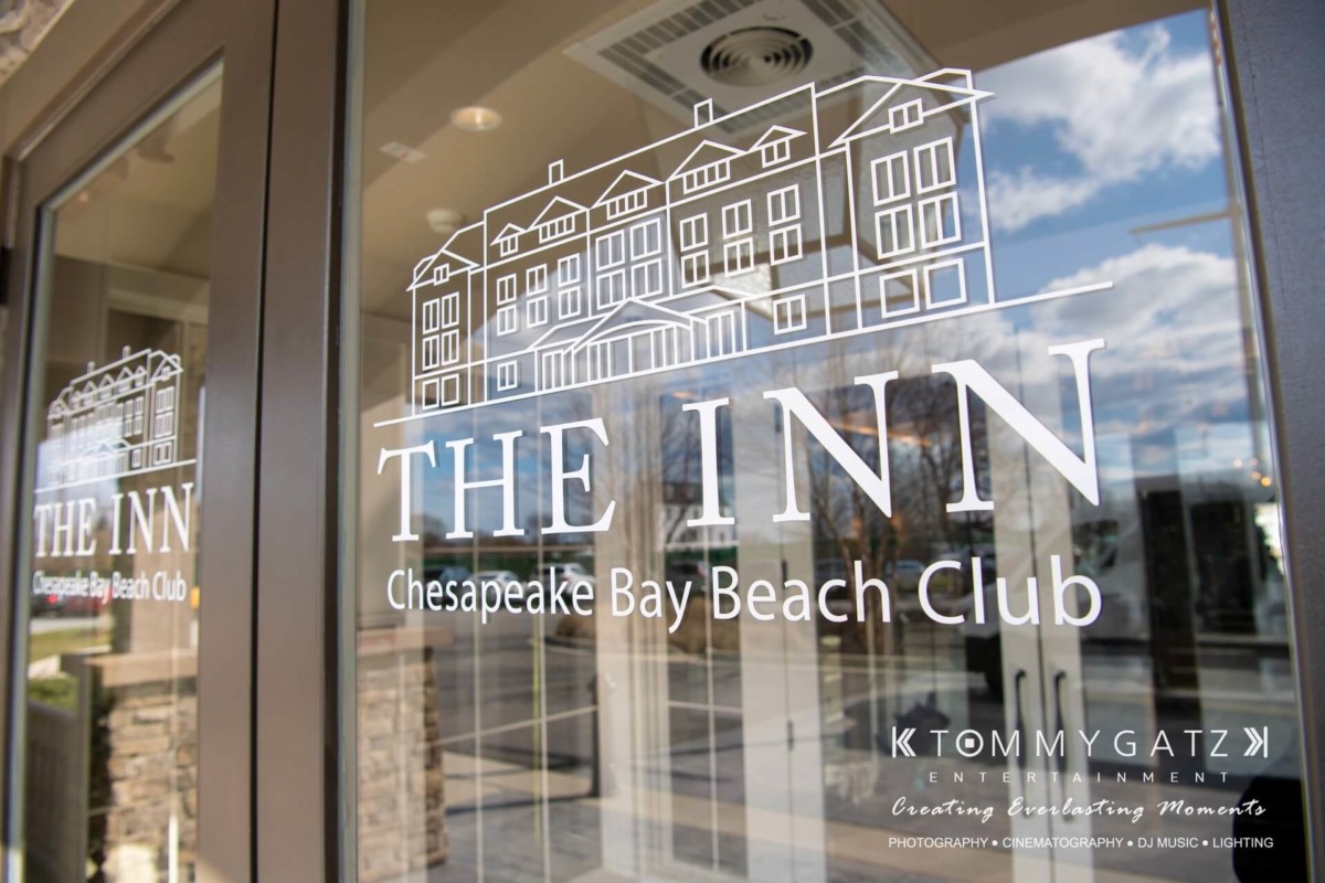Glass entrance doors of The Inn at Chesapeake Bay Beach Club, featuring a logo with a building illustration and text at the bottom promoting Tommy Gatz Entertainment services.