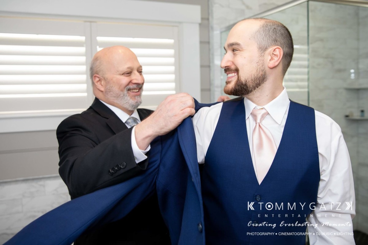 A man helps another man put on a suit jacket in a well-lit room with white shutters and a marble wall.