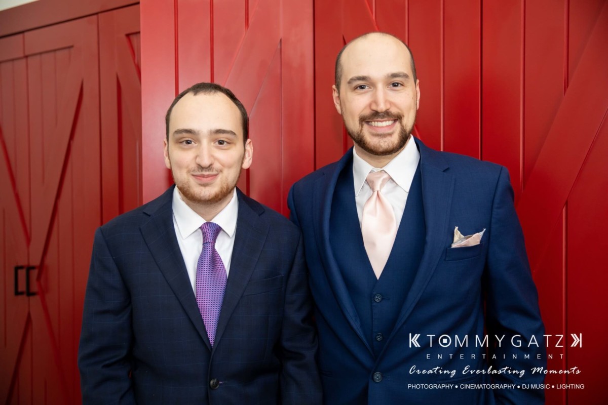 Two men in suits stand in front of a red wall. One is wearing a dark blue suit with a purple tie, and the other a lighter blue suit with a pink tie.