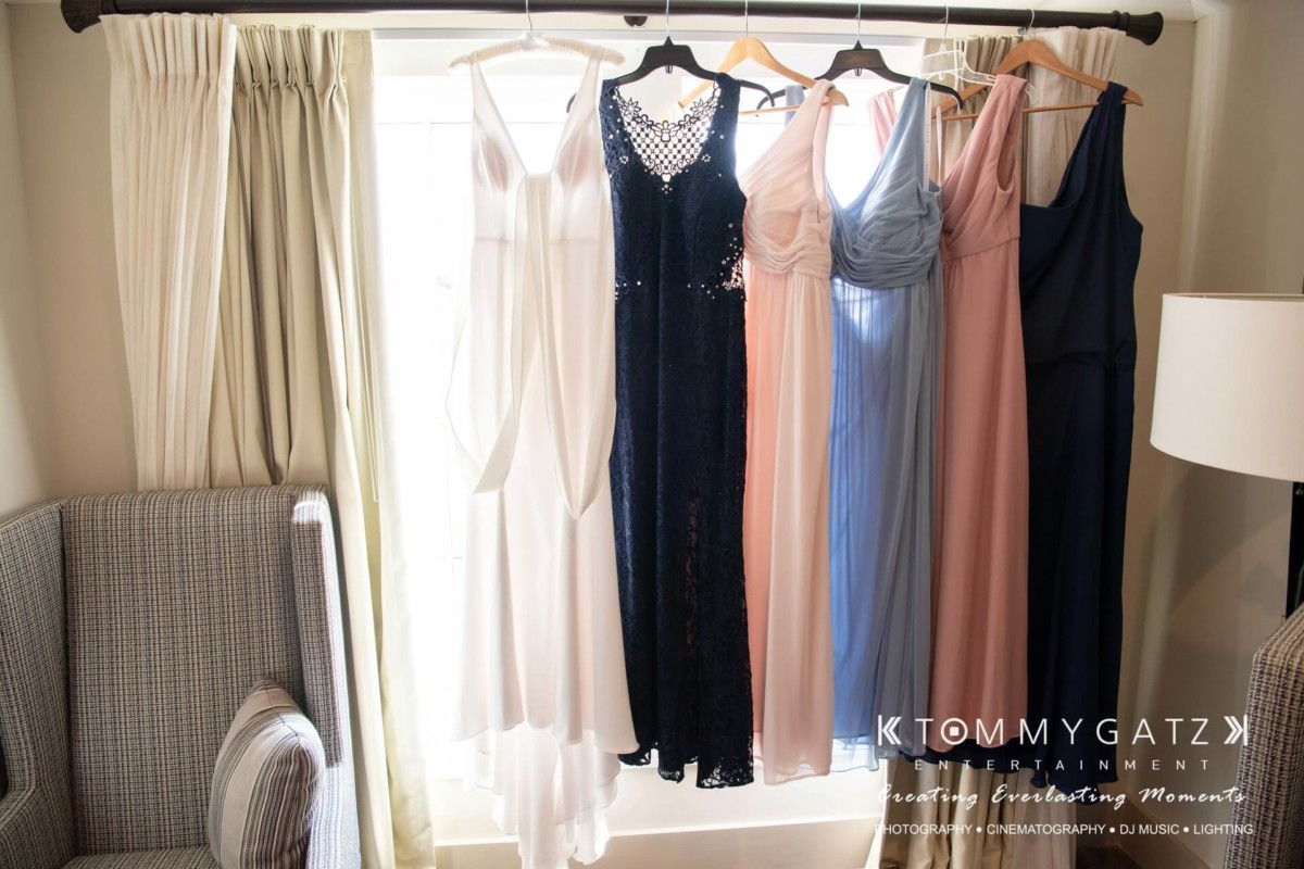 Five dresses in white, navy, pink, and blue hang on a curtain rod in a brightly lit room next to an armchair and lamp.