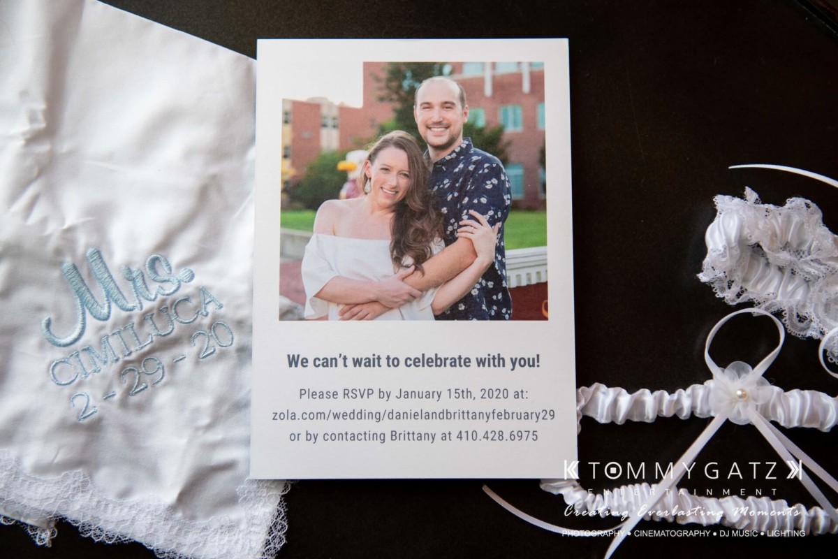 Wedding invitation with a smiling couple's photo. Text includes RSVP instructions and date "2-29-20." Embroidered handkerchief reads "Mr. & Mrs. Cimilluca 2-29-20" beside a garter.