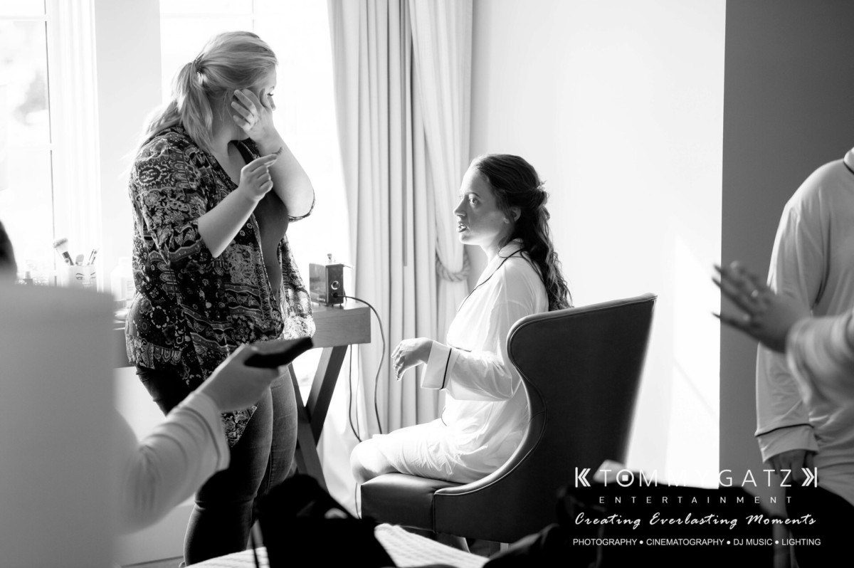 A bride sits in a chair, getting ready as a stylist stands nearby. Another person is partially visible in the foreground.