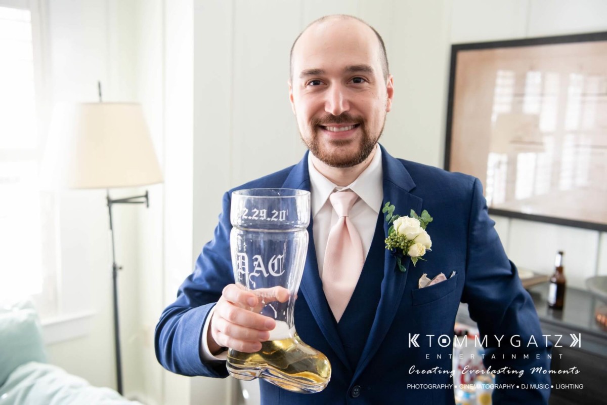 A man in a suit holds a clear boot-shaped glass with liquid, standing indoors. A boutonniere is on his lapel, and "2.29.20" is etched on the glass.