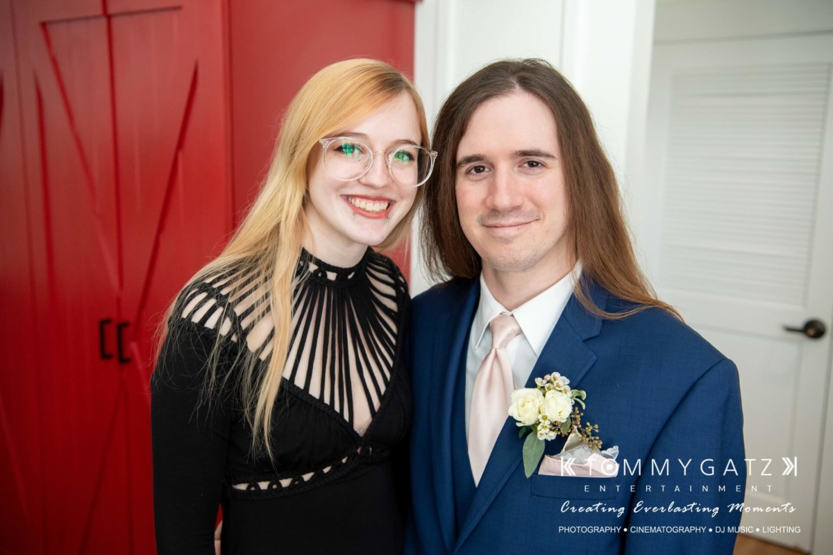 A woman in a black dress and a man in a blue suit with a boutonniere stand smiling in front of red double doors.
