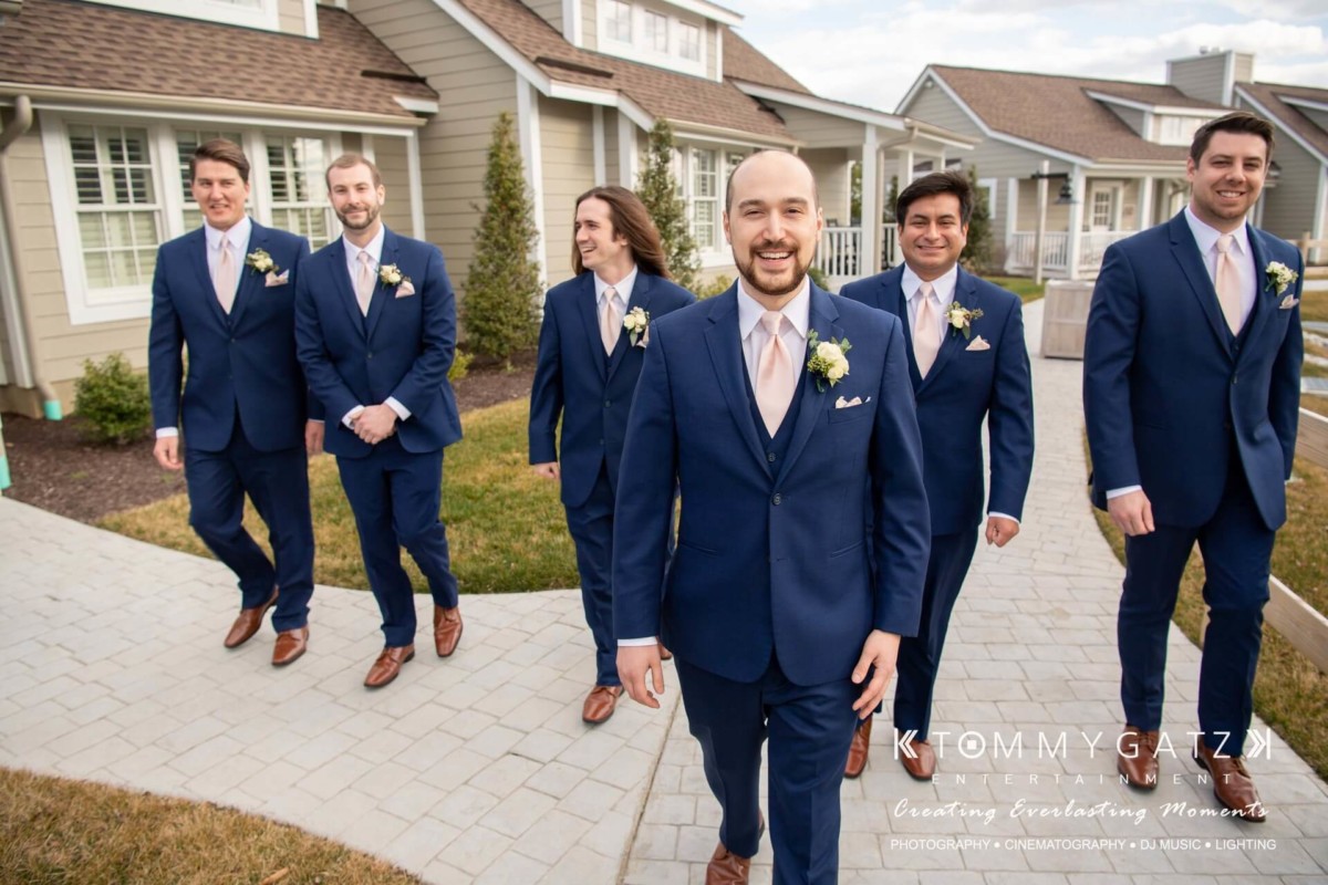A groom and five groomsmen in matching blue suits and pink ties walk outside on a stone path near houses.