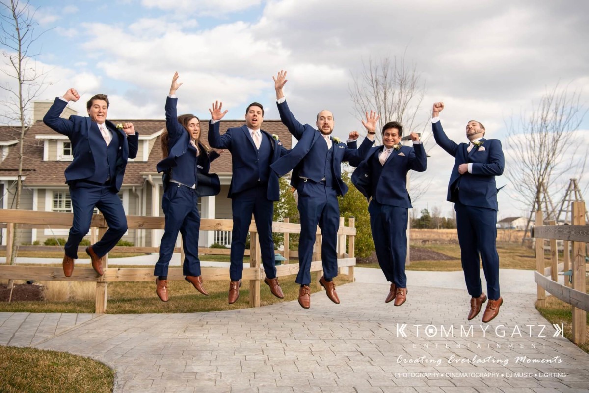 Six men in blue suits jump joyfully on a paved pathway in front of a house, with a clear sky and trees in the background.