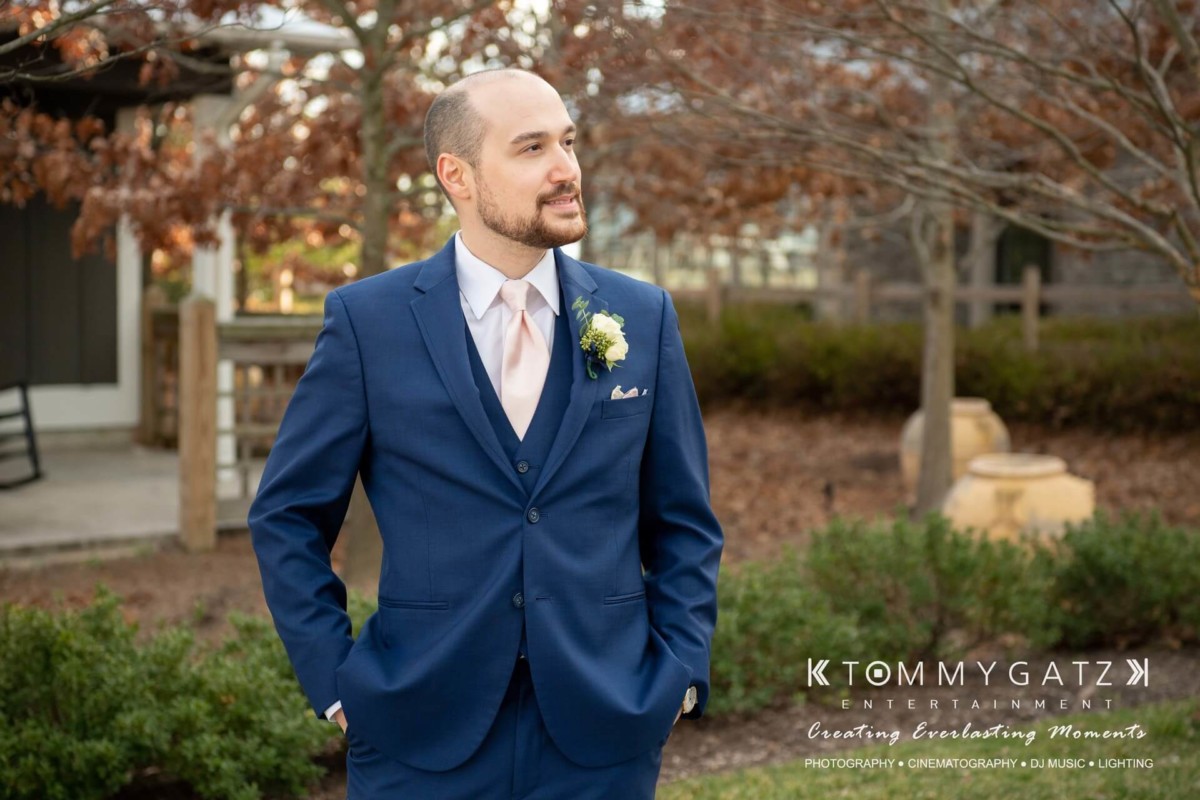 A man in a blue suit with a boutonniere stands outdoors, looking to the side. Behind him are trees and a wooden fence. The image has a logo for Tommy Gatz Entertainment.