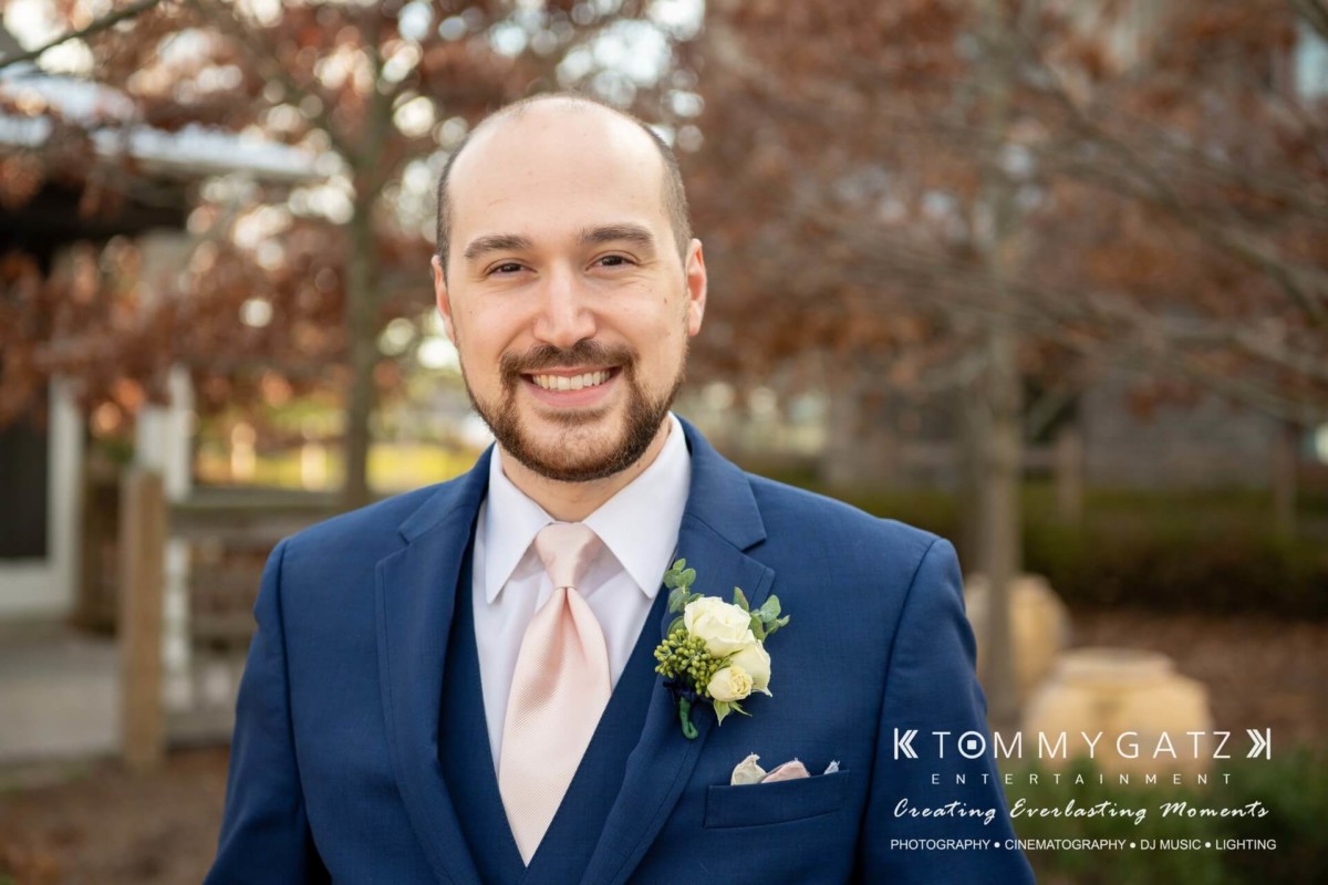 A man in a blue suit with a pink tie and boutonniere smiles outdoors. The background shows blurred trees and a building.