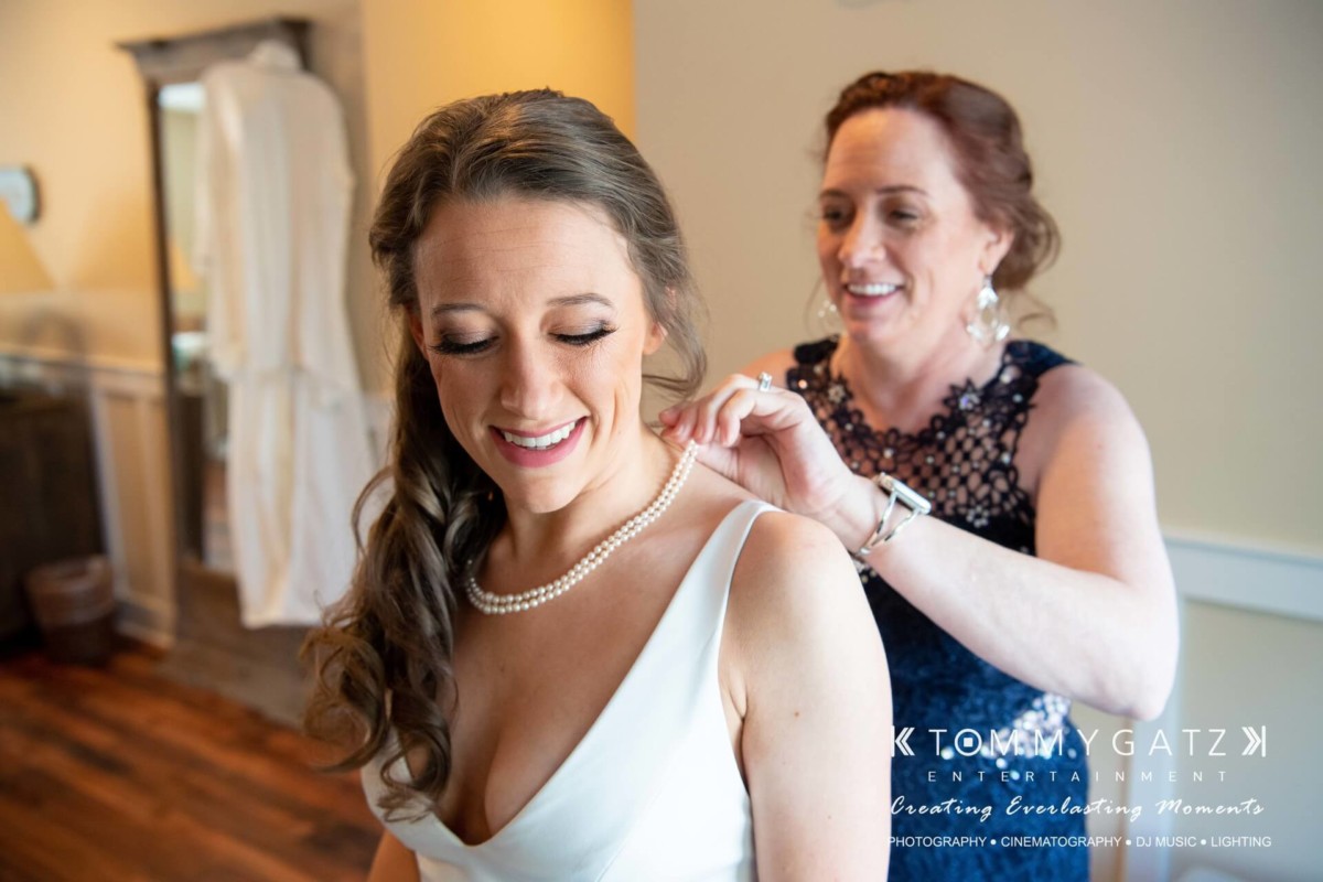 A woman helps another woman put on a necklace in a well-lit room. They both appear happy.