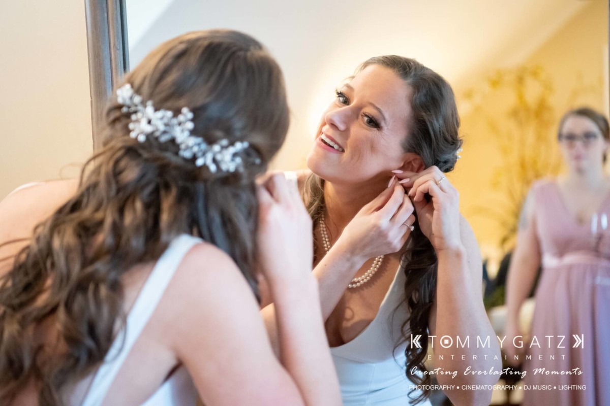 A woman in a white dress is adjusting an earring while looking into a mirror. Another woman in a pink dress is visible in the background.