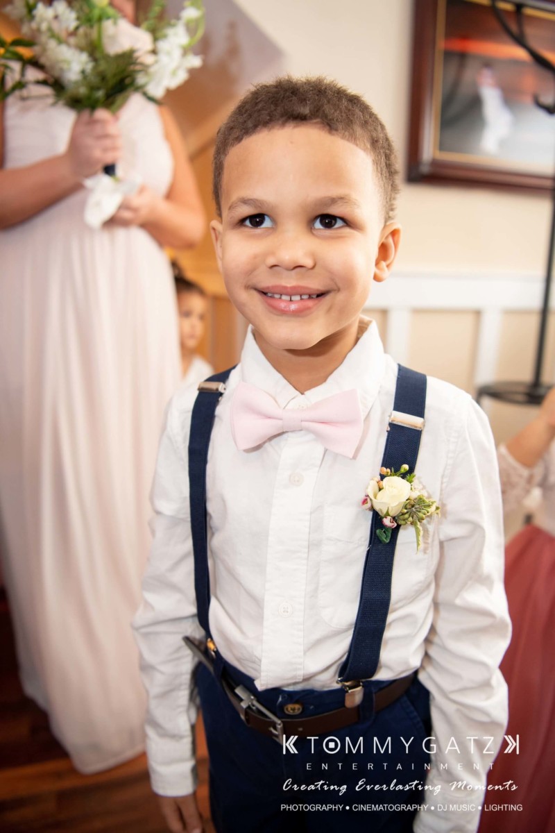 A smiling boy in a white shirt, pink bow tie, and navy suspenders stands indoors. He is wearing a boutonniere. A person in the background holds flowers.
