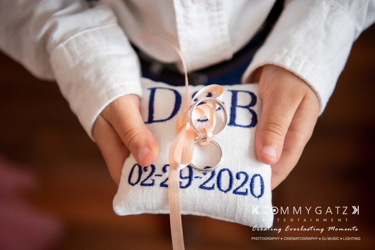 Child holding a white pillow with rings tied by a ribbon, embroidered with initials "D & B" and date "02-29-2020".