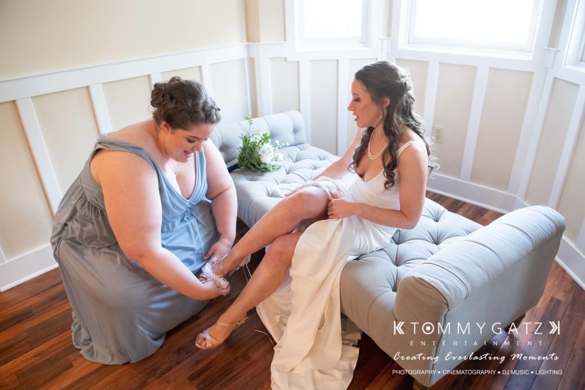 A bridesmaid helps a bride adjust her shoe while sitting on a gray couch in a well-lit room.