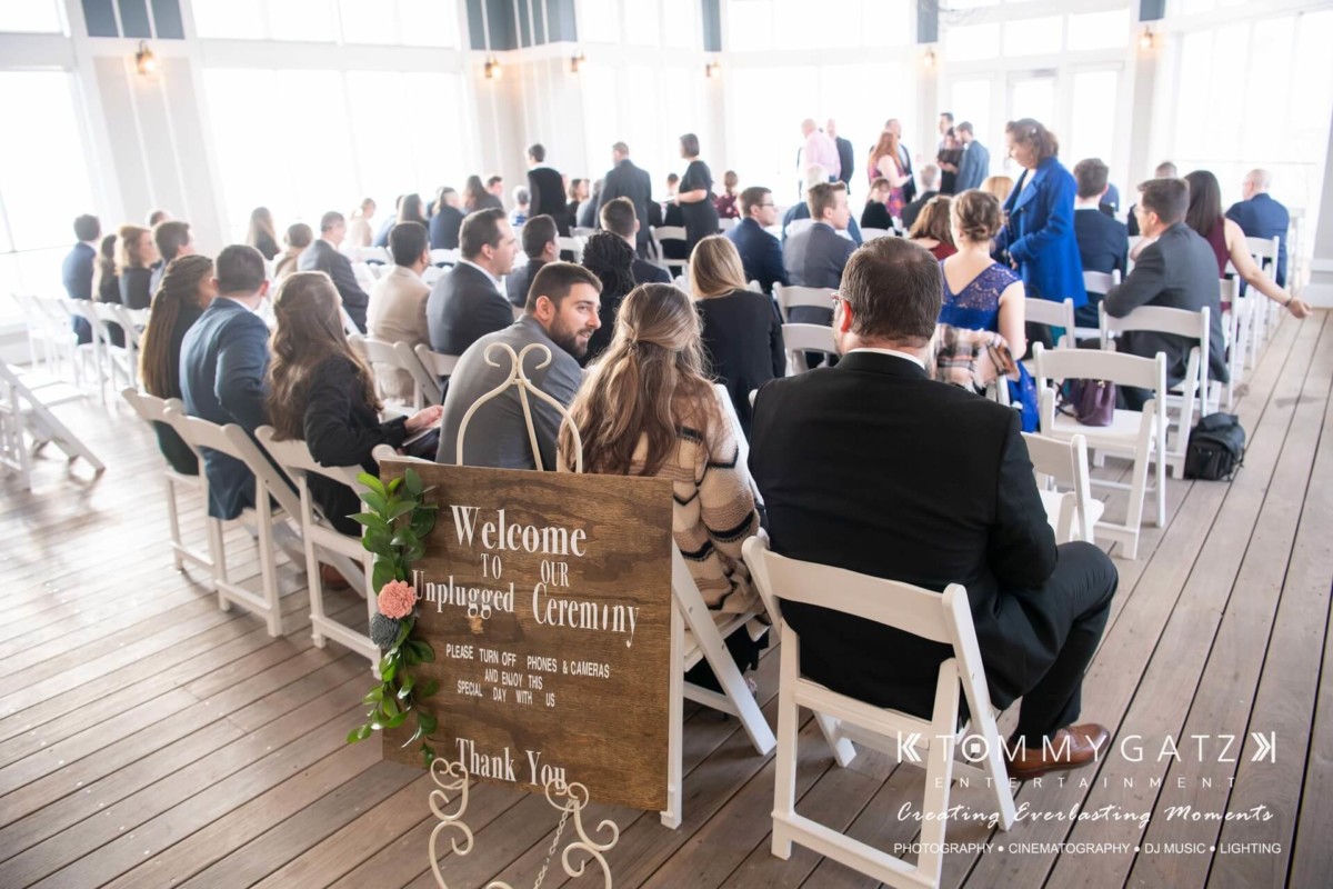 Guests seated indoors for a wedding ceremony, facing a wooden sign that reads "Welcome to our unplugged ceremony.
