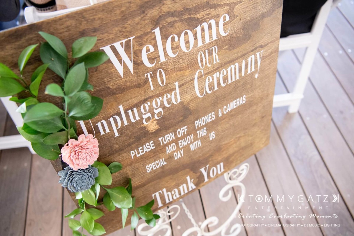 A wooden sign with the text "Welcome to our unplugged ceremony" asking guests to turn off phones and cameras. Decorated with flowers and leaves.