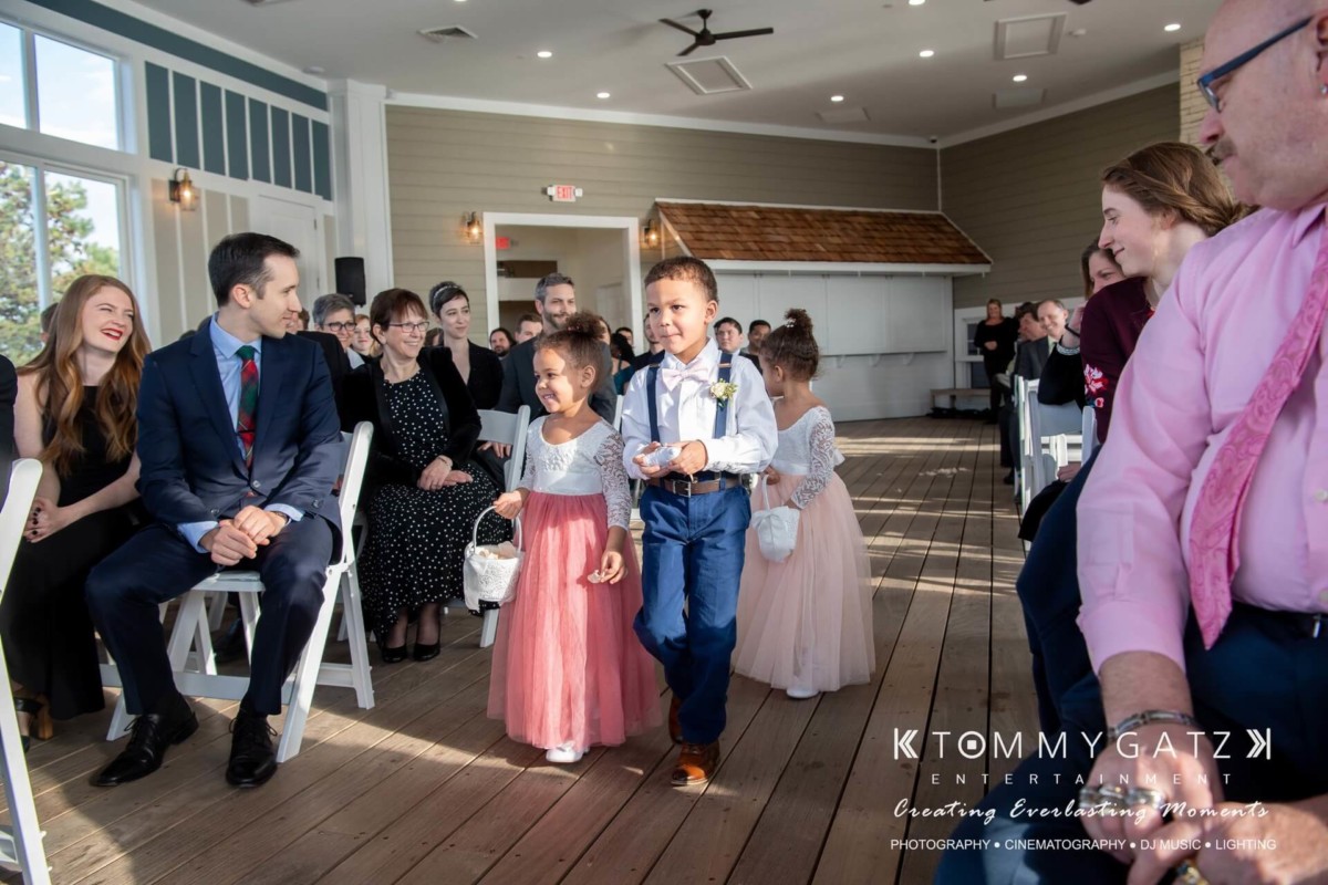 Children dressed formally walk down the aisle carrying flowers at an indoor wedding ceremony, with guests seated on either side.