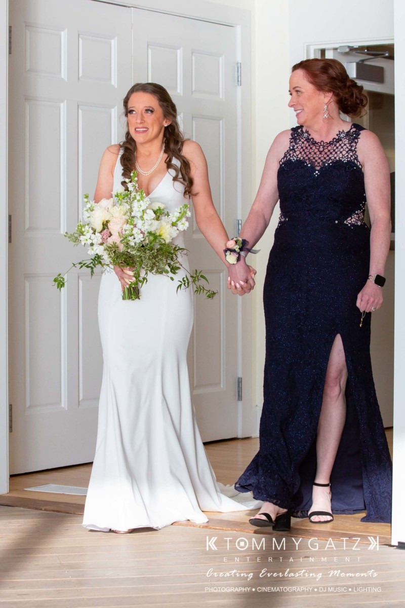 A bride in a white dress and a woman in a navy gown holding a bouquet walk arm-in-arm through a doorway.