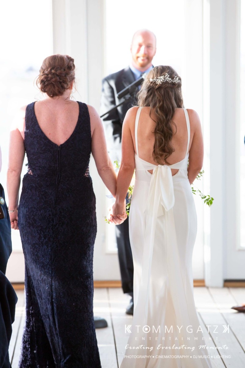Two women, one in a dark blue dress and the other in a white gown, hold hands and walk towards a man standing in front of a door.