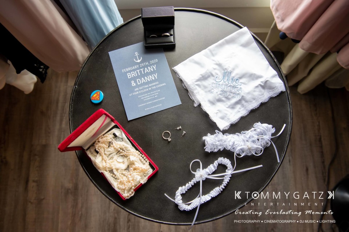 Wedding preparation items on a round table: invitation card, embroidered handkerchief, garter, jewelry, and a ring box.