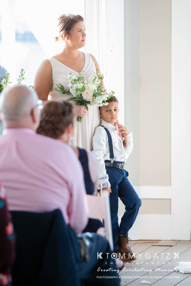 A woman in a white dress holds a bouquet while a boy in a white shirt and blue suspenders stands beside her, next to seated guests.