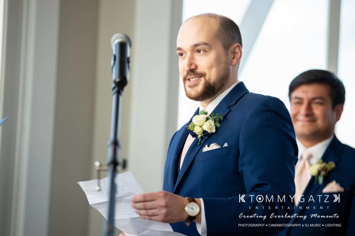 A man in a suit stands at a microphone holding a piece of paper, while another man in a suit stands smiling in the background.