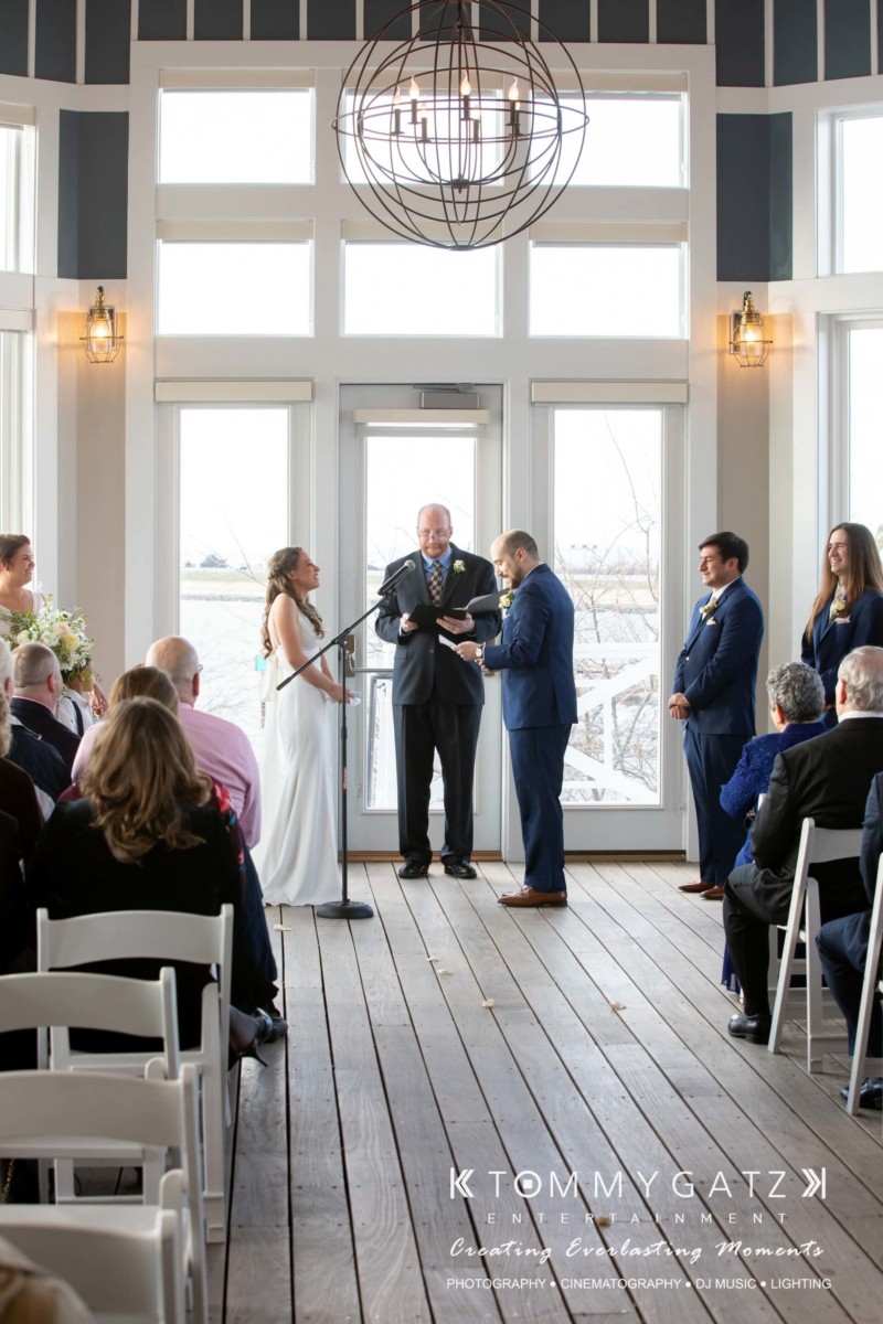 A wedding ceremony with a bride and groom standing in front of an officiant, surrounded by guests seated on white chairs in a bright room.