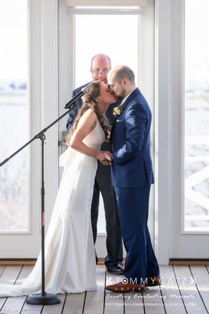A couple is sharing a kiss during their wedding ceremony, with a man standing behind them smiling, in front of a bright window.