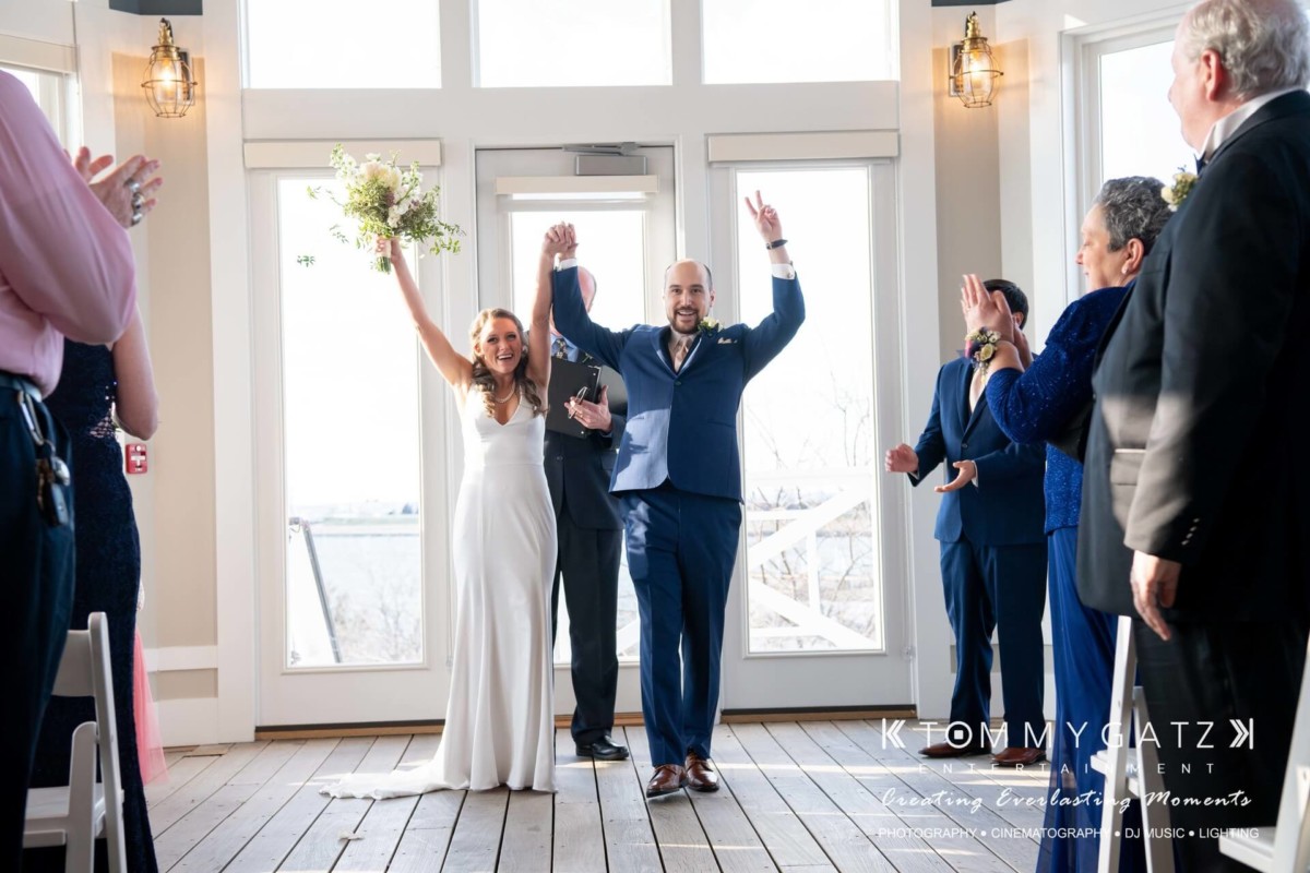 A newlywed couple celebrates with raised arms indoors, surrounded by clapping guests.