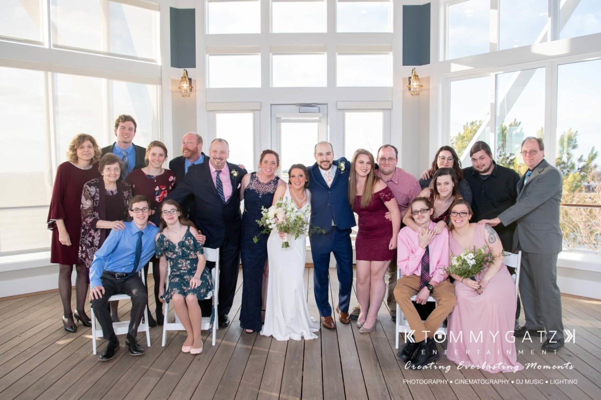 A wedding group photo with a couple in the center, surrounded by family and friends on a wooden deck. Some hold flowers, and everyone is dressed in formal attire.