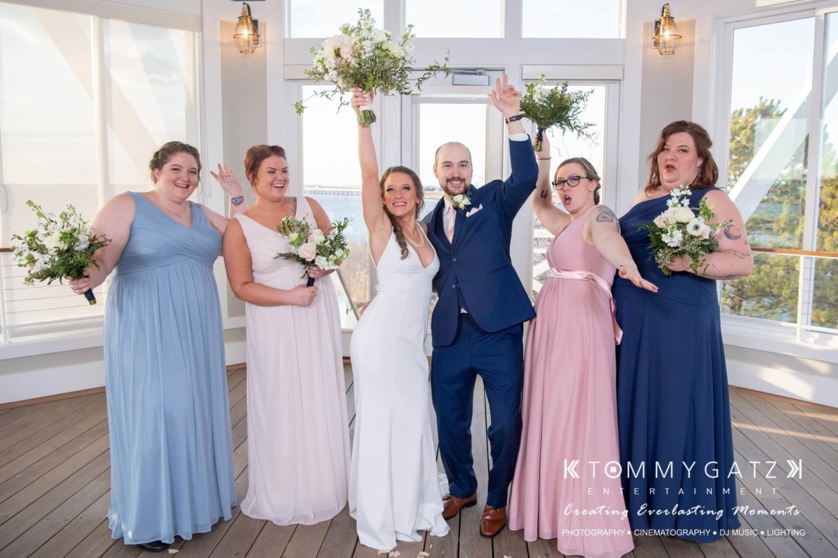 A wedding couple and four bridesmaids pose cheerfully inside a bright room with bouquets and celebratory gestures.