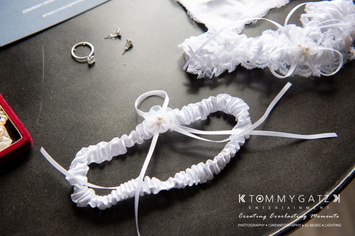 White bridal garter with ribbons and lace on a table, accompanied by earrings and rings.