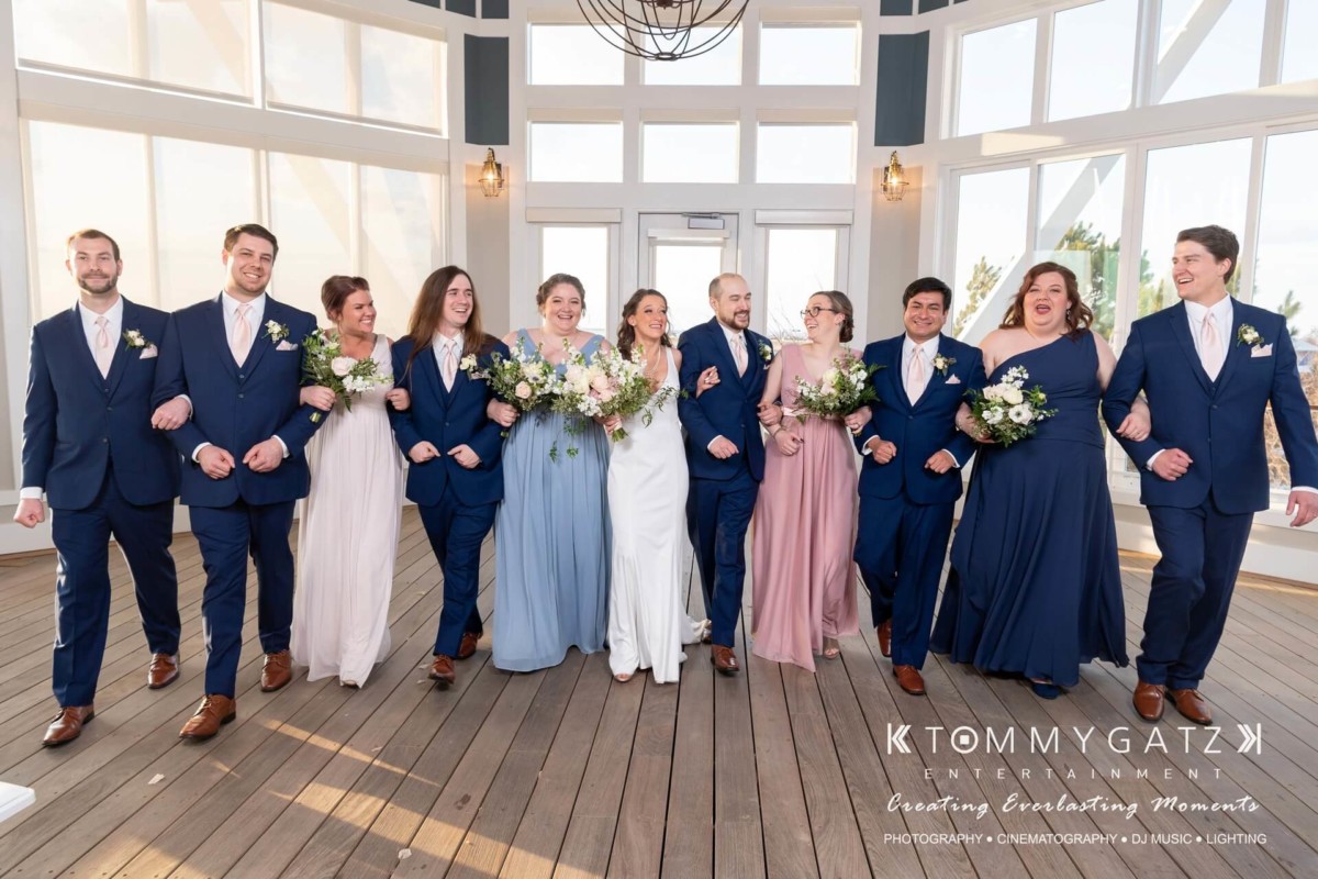 A wedding party walks arm-in-arm indoors. The bridesmaids wear pastel dresses, and the groomsmen wear navy suits with pink ties. The central couple wears a white dress and navy suit.