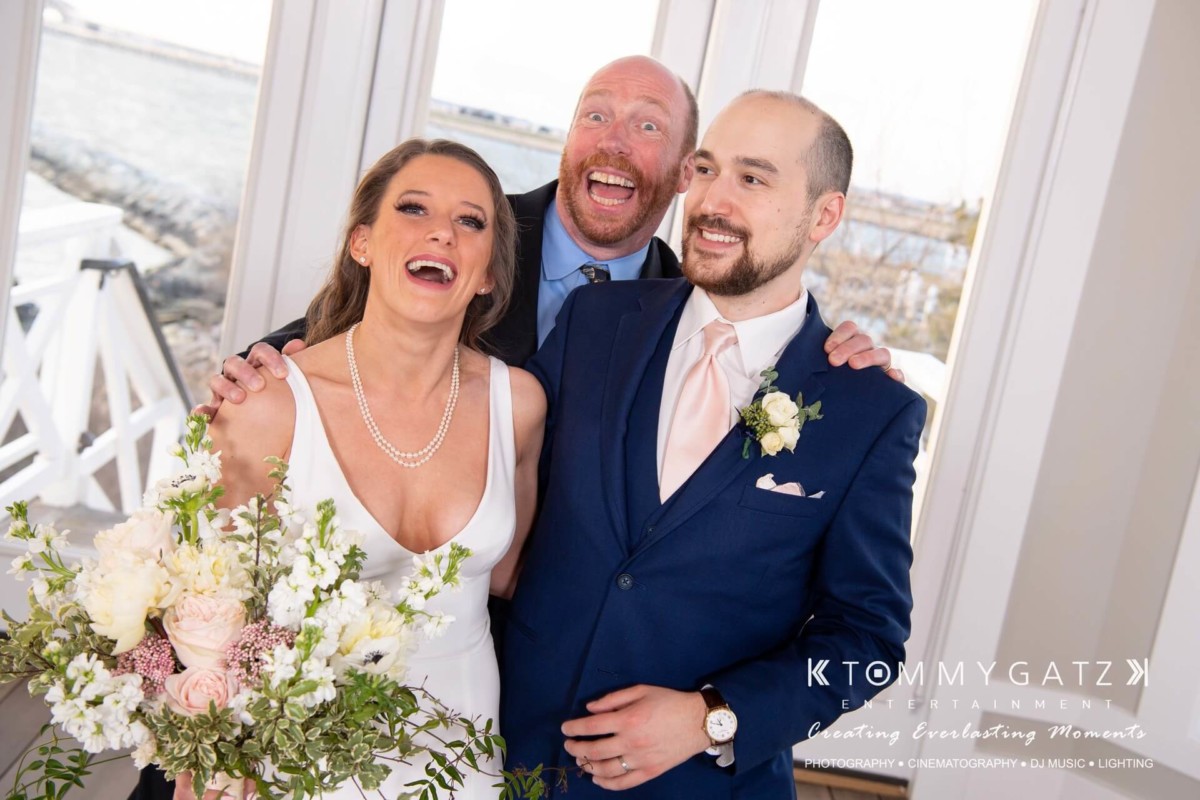 A newlywed couple poses with a smiling man indoors. The bride holds a bouquet of flowers.