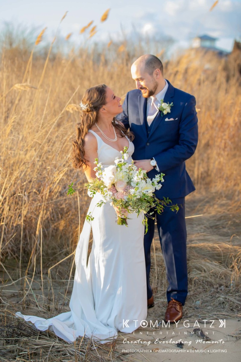A couple stands together outdoors. The woman holds a bouquet and wears a white dress, while the man is in a blue suit. They are surrounded by tall grass under a clear sky.