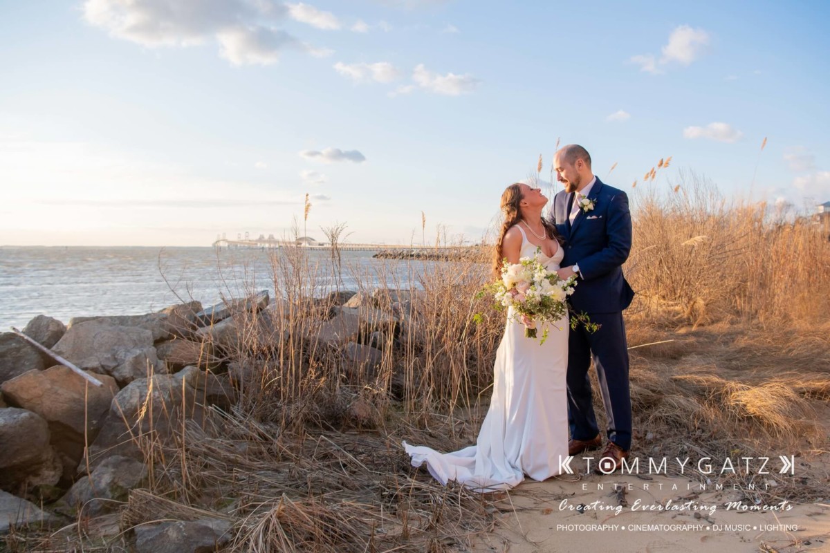 A couple in wedding attire stands on a grassy shoreline, embracing by the water under a partly cloudy sky.