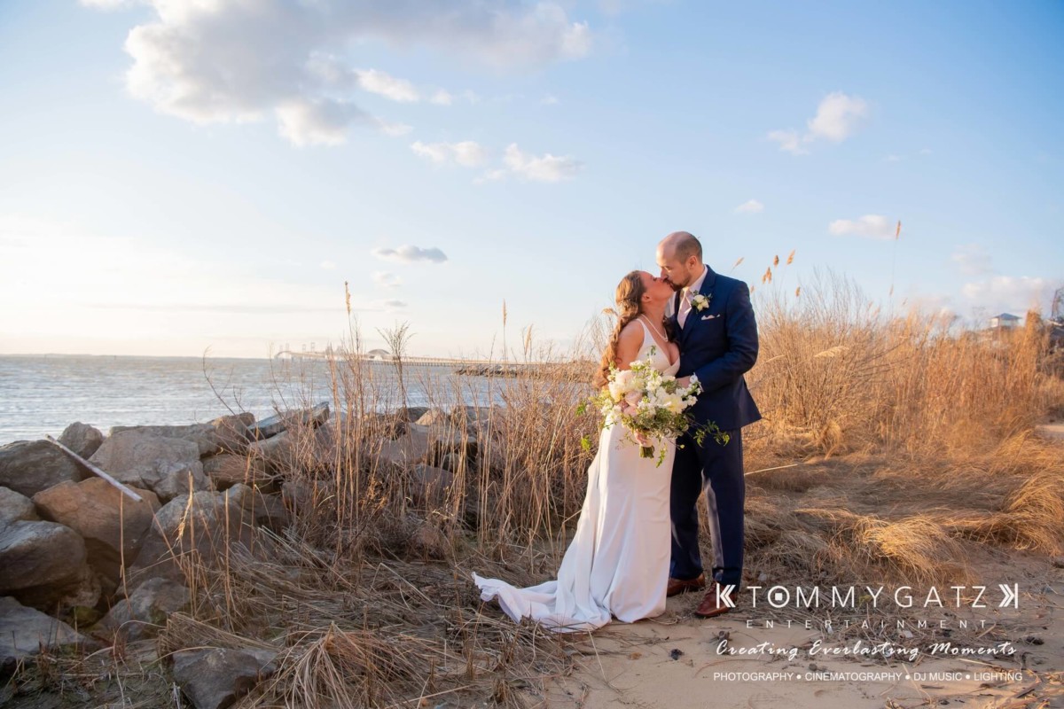Wedding couple embracing near a rocky shoreline with tall grass and a calm sea under a clear sky.
