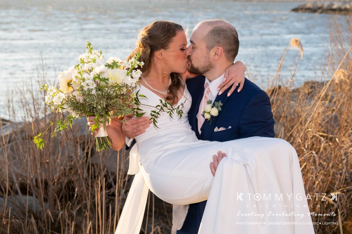 A bride and groom share a kiss by the water. The groom is wearing a navy suit and carrying the bride, who is holding a bouquet of white flowers.